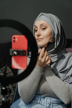 A woman in a hijab applies makeup while filming a beauty tutorial indoors.