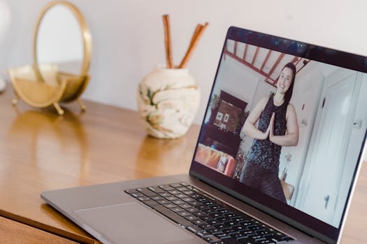 A woman participates in an online yoga class, seen on a laptop screen at home.