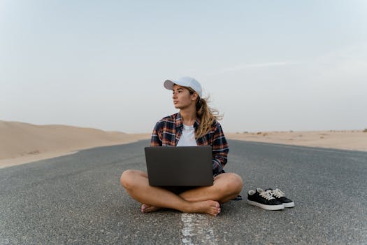 A woman works remotely on a laptop while sitting barefoot on a desert road.