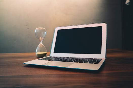 Modern laptop on a wooden desk beside an hourglass, symbolizing time and technology intersection.