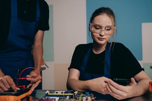 Two young engineers working with electronics, using smartphones and safety glasses for protection.