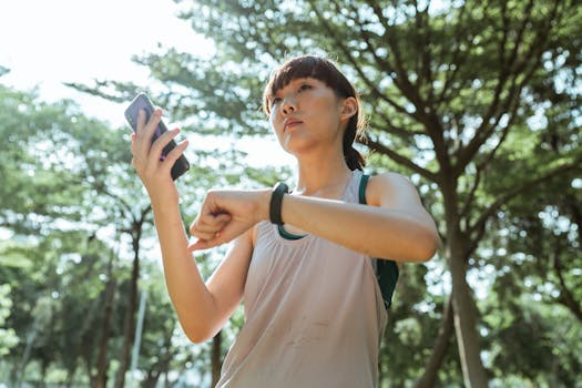 Young woman in sportswear checks smartwatch while using smartphone outdoors in a sunny park.