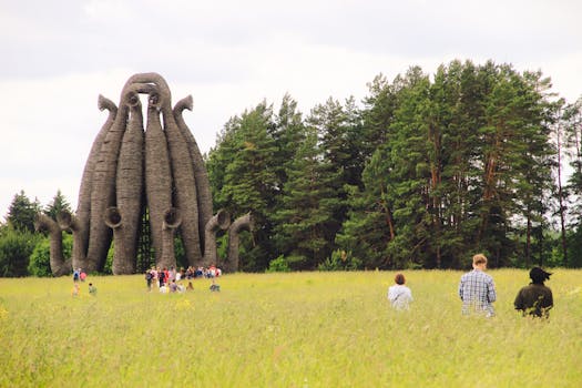 A group of people exploring a unique tower sculpture amid a vibrant green field and forest backdrop.