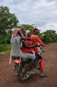 A group of people riding a motorcycle on a dirt road in a rural area surrounded by greenery.