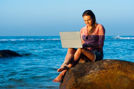 A woman relaxes on a rock by the sea in Sri Lanka, using her laptop during a summer day.