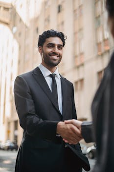 Confident businessman in black suit smiling while shaking hands outdoors in a city.