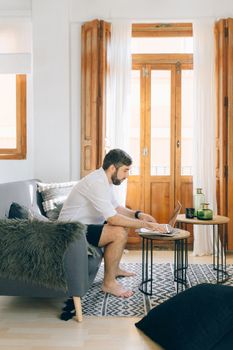 Caucasian man working on a laptop at home, sitting on a couch, with natural light illuminating the room.