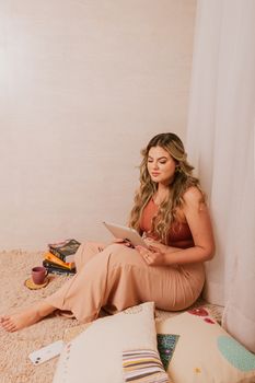 Woman relaxing and reading on a tablet with coffee and books by her side in a cozy room.